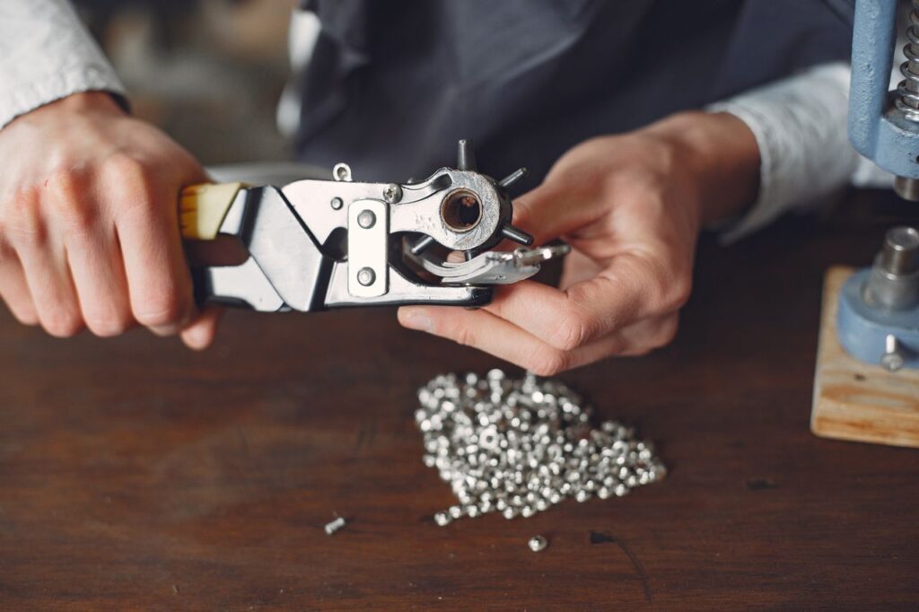 Crop anonymous craftsman in apron using revolving hole punch machine to make holes in leather strap while working in professional workshop